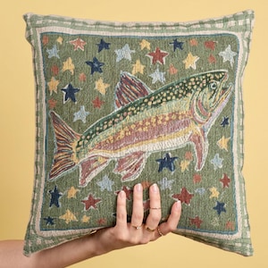 May include: A square woven throw pillow featuring a central trout fish graphic against a sage green background filled with small multi-colored stars. The pillow has a decorative border with a light green and cream checkered pattern.