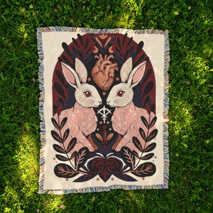 May include: A cream-coloured throw blanket with a decorative design featuring two pink rabbits facing each other, a brown heart, and botanical elements. The blanket has a fringed edge and is set against a green grass background.