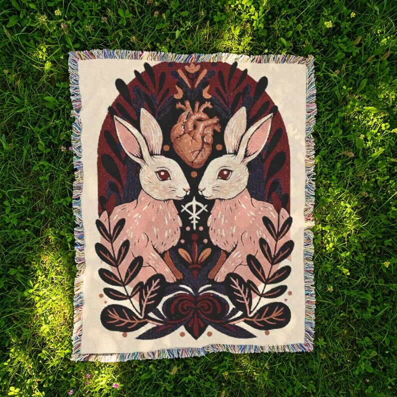 May include: A cream-coloured throw blanket with a decorative design featuring two pink rabbits facing each other, a brown heart, and botanical elements. The blanket has a fringed edge and is set against a green grass background.