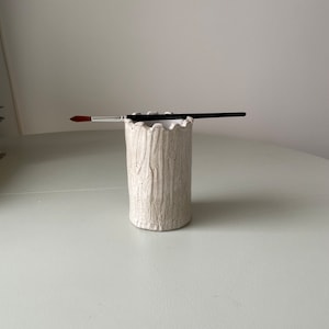May include: A white ceramic brush holder with a wood grain texture. The holder has a scalloped top and is sitting on a light green surface.