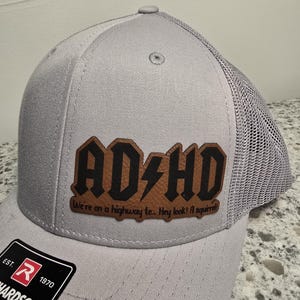 May include: A gray trucker hat with a brown leather patch that says "AD+HD" in black letters with a lightning bolt. The patch also has the text "We're on a highway to... Hey look! A squirrel!" in smaller black letters.