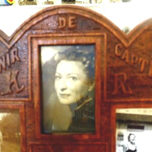 May include: A wooden picture frame with a scalloped edge. The frame has five openings for photos. The top opening has a photo of a woman with dark hair. The frame is labeled "DE SOUVENIR" and "DE CAPTIVITE".