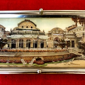 Peut inclure: Un plateau de service rectangulaire argenté avec des poignées. Le plateau présente une image détaillée d'un bâtiment avec un toit en dôme, peut-être un casino ou un théâtre, entouré de jardins et de palmiers. Le plateau est posé sur un fond rouge.