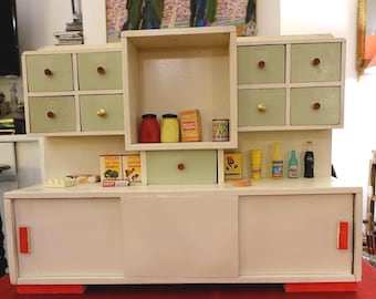 May include: A miniature white and green kitchen cabinet with drawers and a shelf, featuring red and gold knobs. It is filled with tiny food items, including jars, cans, and bottles. The cabinet has a retro design.