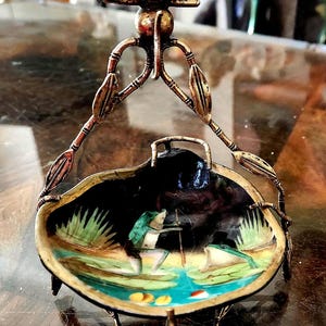Puede incluir: Un plato de metal antiguo y ornamentado con un asa en forma de pájaro. El plato presenta una escena pintada de una persona pescando en un estanque con nenúfares. El plato está sostenido por una base de metal decorativa y tiene un interior oscuro y brillante.
