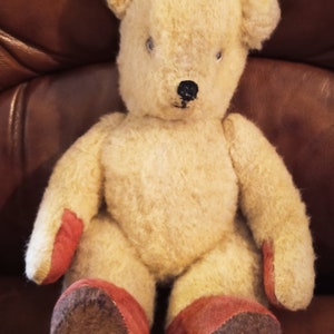 Peut inclure: Un ours en peluche vintage de couleur crème avec des pattes en tissu rouge et marron. L'ours est assis sur un canapé en cuir marron.