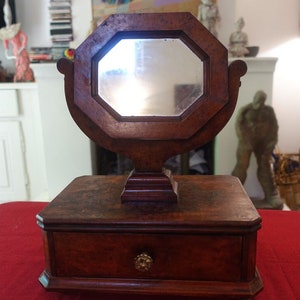 May include: A vintage wooden vanity with an octagonal mirror. The mirror is framed in dark wood and sits atop a small, rectangular base with a drawer. The drawer has a decorative brass knob. The vanity is set on a red surface.