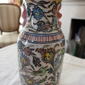 Pode incluir: Um vaso de cerâmica com base branca apresentando padrões florais pintados à mão em azul, amarelo e rosa, juntamente com pequenas ilustrações de pássaros verdes. O vaso tem uma borda alargada, corpo cilíndrico e duas pequenas alças laterais rosas.