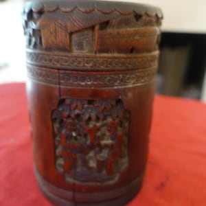Op de afbeelding: Een donkerbruine cilindrische houten container met ingewikkelde houtsnijwerk. De container heeft een deksel en is versierd met een scène van mensen en bomen.