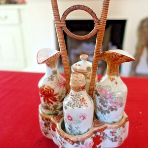 May include: A set of four porcelain condiment bottles with floral designs, held together by a woven wicker stand. The bottles have a white base with gold accents and feature different floral patterns in shades of pink, red, and green.
