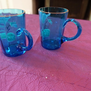 May include: Two blue glass mugs with handles. The mugs have a gold rim and a floral design etched into the glass. The mugs are sitting on a purple surface.