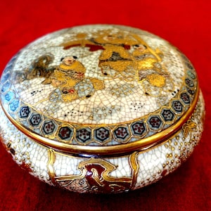 Könnte beinhalten: Runde, antike Schmuckdose aus Keramik mit Deckel. Die Dose hat eine weiße, craquelierte Glasur mit goldenen und blauen Akzenten. Der Deckel ist mit einer goldfarbenen Illustration von Figuren verziert. Die Dose hat einen goldenen Rand.