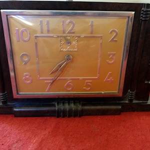 Può includere: Un orologio da tavolo rettangolare con una cassa in finto legno marrone scuro e un quadrante dorato metallizzato. L'orologio presenta numeri argentati da uno a dodici, un bordo rettangolare centrale e due lancette sottili e appuntite.