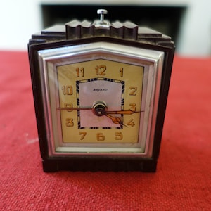 May include: A vintage brown and silver-toned alarm clock with a rectangular face. The clock face is a light tan colour with black numerals and hands. The clock reads "BAJARD" at the centre. The clock is sitting on a red surface.