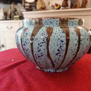 May include: A ceramic planter with a light blue speckled glaze and vertical brown drip pattern. The round planter has a wide opening and sits on a red surface. Decorative pottery for indoor or outdoor use.