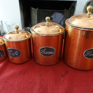May include: Set of four copper canisters with lids. Each canister has a label with a different word: "Thé", "Café", "Sucre", and "Farine". The canisters are cylindrical with a shiny copper finish and gold-colored lids.