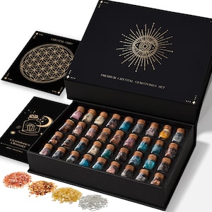 May include: A black box set with a gold design and the text "Premium Crystal Gemstones Set" contains small glass bottles filled with colorful gemstones. Two black cards with gold designs are next to the box. Several piles of gemstones are in the foreground.