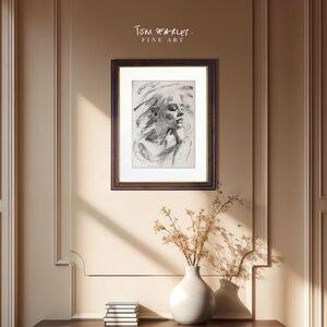 Puede incluir: Un retrato en blanco y negro enmarcado de una mujer con el pelo suelto. La obra de arte se muestra en un marco de madera marr&oacute;n oscuro con un passe-partout blanco. El texto "Tom Searles. Fine Art" est&aacute; en la esquina superior derecha. Un jarr&oacute;n blanco con flores secas est&aacute; sobre una mesa de madera oscura.