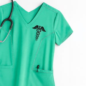 May include: A teal green medical scrub top with a V-neck and a black embroidered caduceus symbol on the chest. A black stethoscope hangs on the left side. A pen is in the pocket.