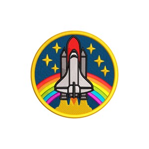 Puede incluir: Parche bordado con un diseño de transbordador espacial. El parche circular tiene un borde amarillo. El transbordador es blanco y gris con detalles en rojo, sobre un arcoíris y llamas amarillas. El fondo es azul oscuro con estrellas amarillas.