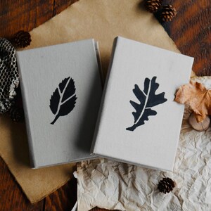May include: Two small, gray, fabric-covered journals with black leaf designs. The journal on the left has a simple leaf design, while the journal on the right has an oak leaf design.