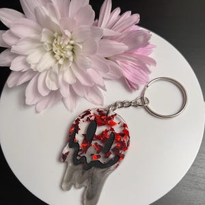May include: A red and white resin keychain with a dripping smiley face design. The keychain has red glitter embedded in the resin.