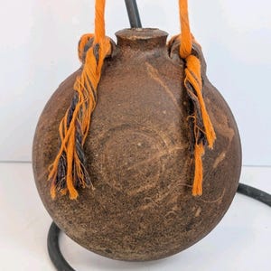 May include: A brown, round ceramic flask with a narrow neck and orange and black rope handles. The flask has a textured surface and appears to be handmade. The rope is knotted at the top and hangs down on either side of the flask.