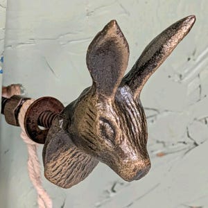May include: A bronze-toned metal cabinet knob shaped like a rabbit head with long ears, textured fur details, and closed eyes. The knob includes a threaded metal bolt, a circular washer, and a nut, with a piece of light-colored string attached.
