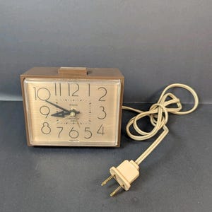 May include: A brown rectangular Westclox quartz alarm clock with a light brown face and a clear plastic cover. The clock has black hands and numbers. The power cord and plug are beige.