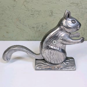 May include: A silver-colored metal nutcracker in the shape of a squirrel. The squirrel is standing on a rectangular base, with its tail forming a lever. The nutcracker is designed to crack nuts with its mouth.