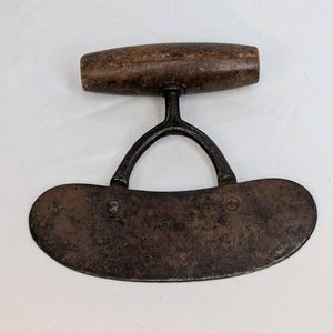 May include: A vintage herb chopper with a curved, rusty blade and a wooden handle. The handle is a dark brown color and is attached to the blade with a curved metal support. The blade is secured with two rivets.