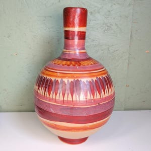 May include: A round ceramic vase with a narrow neck, featuring horizontal bands of red, orange, and purple paint. The body is decorated with a repeating pattern of vertical, petal-like shapes in shades of red and purple against a light tan base.