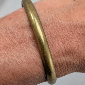 May include: A solid, smooth, cylindrical brass bangle bracelet with a polished metallic finish. The bracelet features a continuous, seamless circular band design that rests against the skin, showcasing a warm, muted gold-toned metal surface.