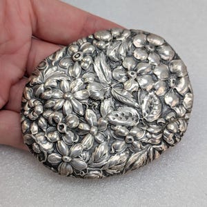 May include: An oval-shaped silver belt buckle featuring a highly detailed, raised relief design of various flowers and leaves covering the entire surface. The metallic finish has a polished sheen with darkened recessed areas to highlight the floral pattern.