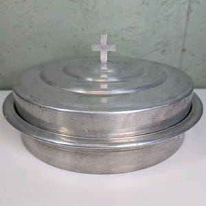 May include: A round, silver-colored metal container with a flat base and a matching lid. The lid features a small, centered cross-shaped handle on top. The surface of the metal has a brushed finish with concentric circular indentations on the lid.