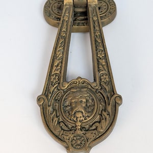 May include: An antique bronze door knocker with an ornate design. The knocker features a central face within a circular frame, surrounded by floral and scrollwork details. The knocker is a warm, aged bronze color.