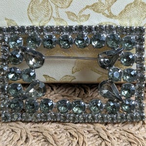 May include: A rectangular, silver-toned buckle with rows of small, clear rhinestones and larger, smoky-gray stones. The buckle has a pin closure and a decorative, open center. The stones are set in a symmetrical pattern, creating a vintage or antique aesthetic.