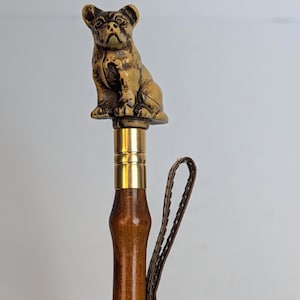 May include: A decorative walking cane with a detailed, golden-colored bulldog figurine atop a wooden handle. The cane features a brass collar and a brown leather wrist strap, offering both style and support. The overall design is classic and elegant.