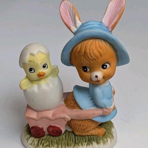 May include: A ceramic Easter figurine featuring a bunny wearing a blue hat and dress, pulling a pink wagon with a yellow chick hatching from an egg. The figurine is set on a base of green grass.