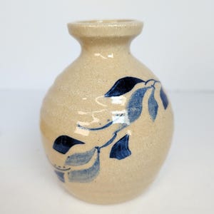 May include: A small, beige ceramic vase with a rounded body and a narrow neck. The vase is decorated with a blue leaf design. The vase is approximately 5 inches tall.