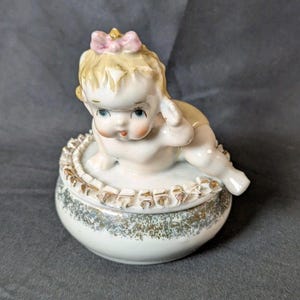 May include: A vintage porcelain trinket box featuring a crawling baby figurine with a pink bow. The box is white with a gold and gray speckled design and a gold trim.