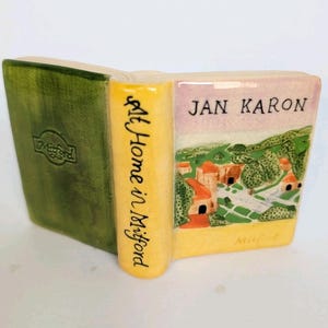 May include: A ceramic book figurine with a green cover and a yellow spine. The spine reads "At Home in Milford." The book's pages display a landscape scene and the author's name, "Jan Karon."