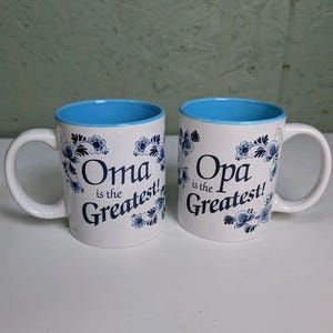 May include: Two white ceramic mugs with light blue interiors and handles. Each mug features dark blue floral patterns and text. One mug reads "Oma is the Greatest!" and the other reads "Opa is the Greatest!" in a dark blue serif font.