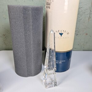 May include: A clear, faceted crystal obelisk with etched designs, standing in front of a cylindrical Asfour Crystal box with a cream and dark blue label and a separate grey foam protective insert for storage and display purposes.