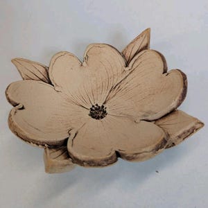 May include: A ceramic flower-shaped dish with a light brown color. The dish has detailed petal and leaf designs, with a dark brown center. The edges are slightly raised, and the overall shape is organic and natural.