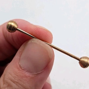 May include: A gold-toned industrial barbell body jewelry piece featuring a straight metal bar with two spherical ends. Each sphere has a textured, ribbed surface design consisting of multiple horizontal grooves circling the round beads.