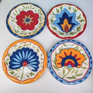 May include: A set of four round ceramic plates featuring hand-painted floral motifs. Each plate displays a unique central flower design in red, blue, or orange, surrounded by green leafy vines and a colorful scalloped border rim.