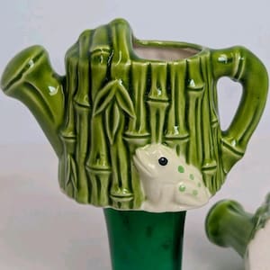 May include: A green ceramic watering can shaped like bamboo, with a white frog figurine on the side. The watering can has a handle and a spout. The frog has green spots and a black eye. The watering can is a decorative item.