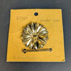 May include: A gold-toned metal scarf clip in a floral design, attached to a tan cardboard backing. The clip is round with petal-like shapes. The cardboard reads "Loree" and "Scarf Clip By Jewelry Concepts".
