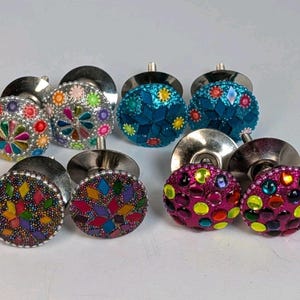May include: A collection of decorative cabinet knobs with colorful, mosaic-style designs. The knobs feature a variety of colors, including silver, blue, pink, green, yellow, and red. Each knob has a metal base and a rounded, jeweled top.